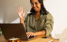 Smiling woman waving at laptop in a sunny room.