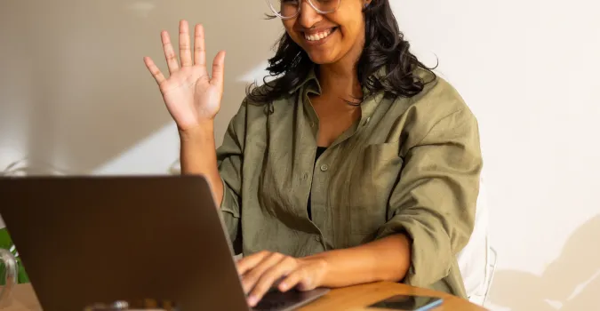 Smiling woman waving at laptop in a sunny room.