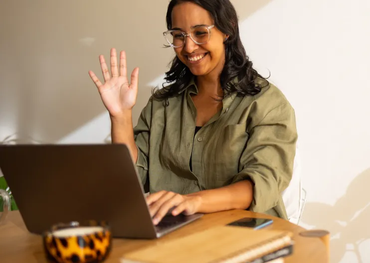 Smiling woman waving at laptop in a sunny room.