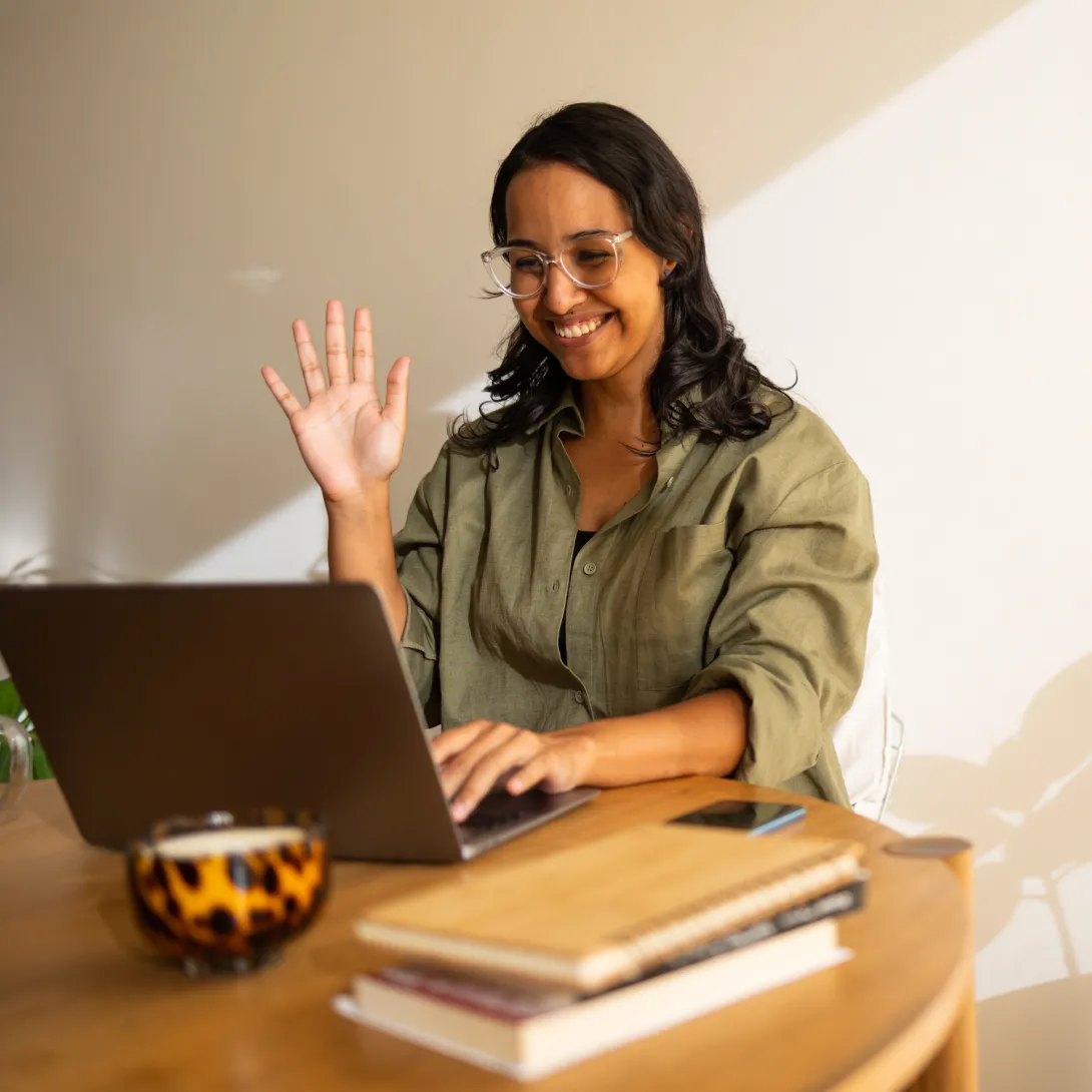 Smiling woman waving at laptop in a sunny room.